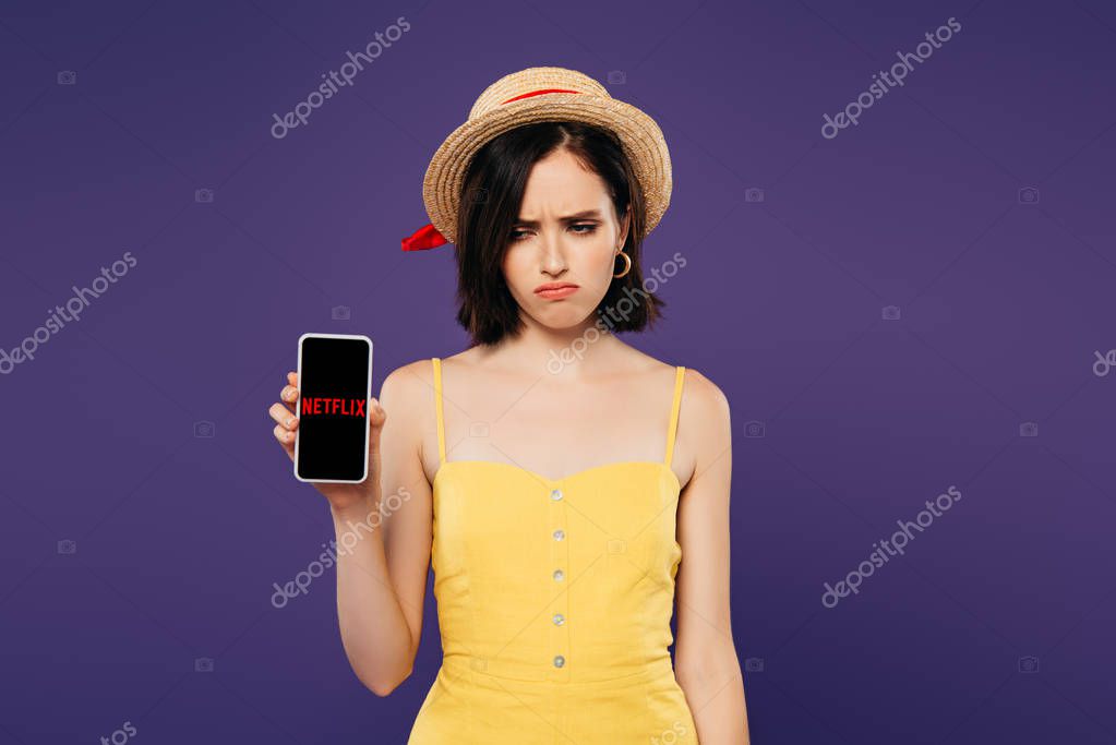 KYIV, UKRAINE - JULY 3, 2019: sad pretty girl in straw hat holding smartphone with netflix app isolated on purple
