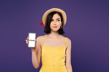 KYIV, UKRAINE - JULY 3, 2019: dreamy pretty girl in straw hat holding smartphone with tinder app isolated on purple  clipart