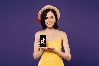 Kiev, Ukrayna - 3 Temmuz 2019: mor izole tiktok uygulaması ile akıllı telefon tutan hasır şapka güzel kız gülümseyen 