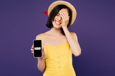 Kiev, Ukrayna - 3 Temmuz 2019: mor izole hbo uygulaması ile akıllı telefon gösteren yüzünde el ile hasır şapka güzel kız gülümseyen 