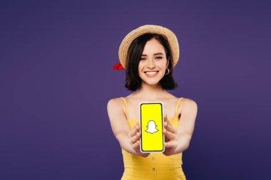 Kiev, Ukrayna - 3 Temmuz 2019: mor izole snapchat uygulaması ile akıllı telefon sunan hasır şapka gülümseyen güzel kız seçici odak 