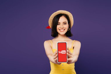 Kiev, Ukrayna - 3 Temmuz 2019: mor izole youtube uygulaması ile akıllı telefon sunan hasır şapka gülümseyen güzel kız seçici odak 