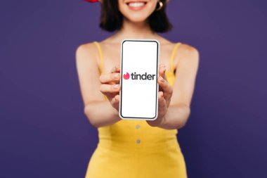 KYIV, UKRAINE - JULY 3, 2019: selective focus of smiling girl presenting smartphone with tinder app isolated on purple  clipart