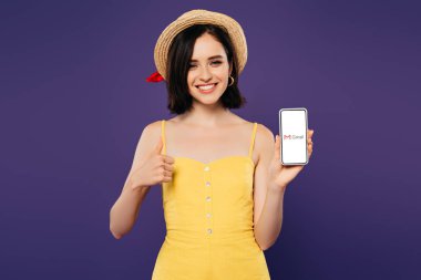 KYIV, UKRAINE - JULY 3, 2019: smiling pretty girl in straw hat showing thumb up while holding smartphone with gmail app isolated on purple  clipart