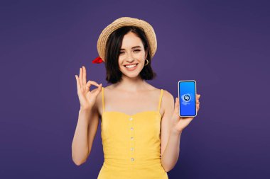Kiev, Ukrayna - Temmuz 3, 2019: Shazam uygulaması ile akıllı telefon tutan hasır şapka güzel kız gülümseyen ve ok işareti mor izole gösteren 