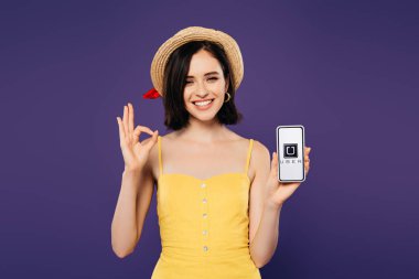 Kiev, Ukrayna - 3 Temmuz 2019: Uber uygulaması ile akıllı telefon tutan ve mor izole ok işareti gösteren hasır şapka güzel kız gülümseyen 