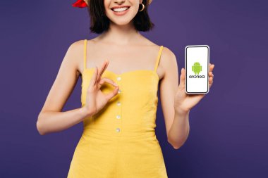 Kiev, Ukrayna - 3 Temmuz 2019: android logolu akıllı telefon tutan ve mor izole ok işareti gösteren gülümseyen kız kırpılmış görünümü 