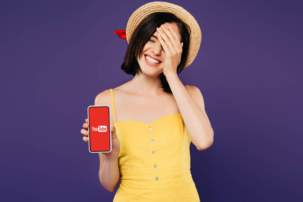 KYIV, UKRAINE - JULY 3, 2019: smiling pretty girl in straw hat with hand on face showing smartphone with youtube app isolated on purple 