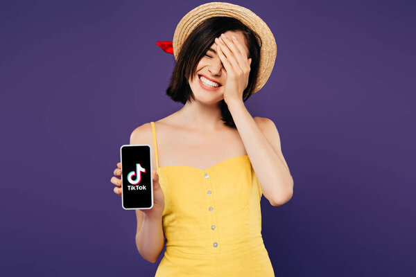 KYIV, UKRAINE - JULY 3, 2019: smiling pretty girl in straw hat with hand on face showing smartphone with tiktok app isolated on purple 
