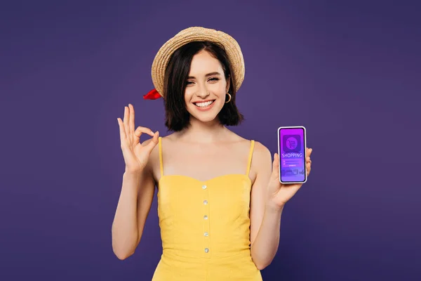 オンラインショッピングアプリでスマートフォンを持ち 紫色に孤立したOkサインを示すわら帽子でかわいい女の子を微笑む — ストック写真