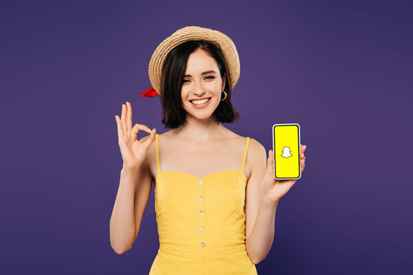 KYIV, UKRAINE - JULY 3, 2019: smiling pretty girl in straw hat holding smartphone with Snapchat app and showing ok sign isolated on purple 