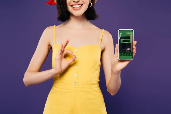 裁剪视图微笑的女孩在拿着智能手机与预订应用程序 并显示确定标志隔离在紫色 — 图库照片