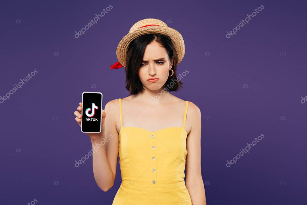 KYIV, UKRAINE - JULY 3, 2019: sad pretty girl in straw hat holding smartphone with tiktok app isolated on purple