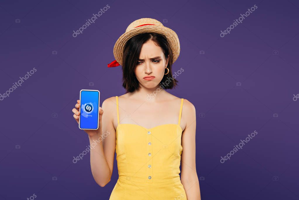 KYIV, UKRAINE - JULY 3, 2019: sad pretty girl in straw hat holding smartphone with shazam app isolated on purple