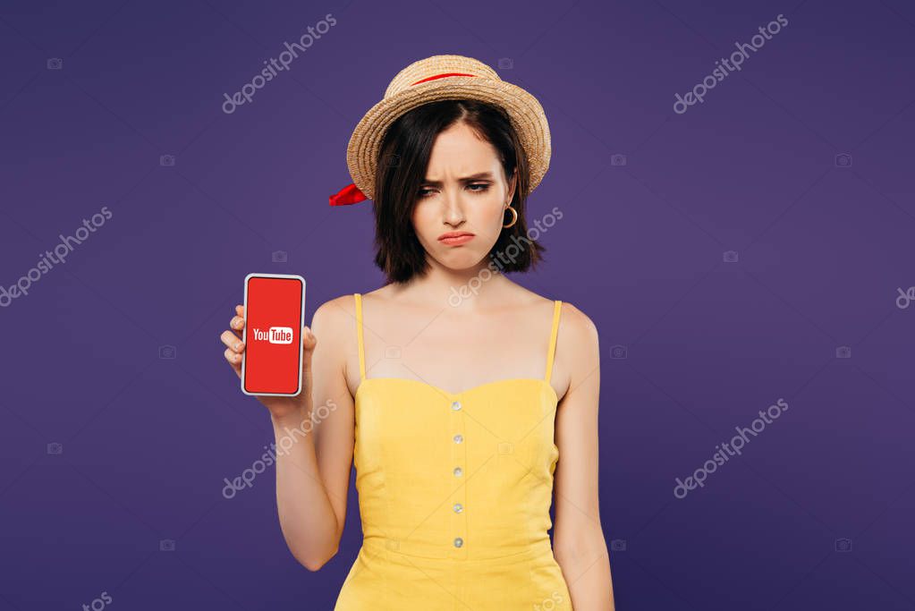 KYIV, UKRAINE - JULY 3, 2019: sad pretty girl in straw hat holding smartphone with youtube app isolated on purple
