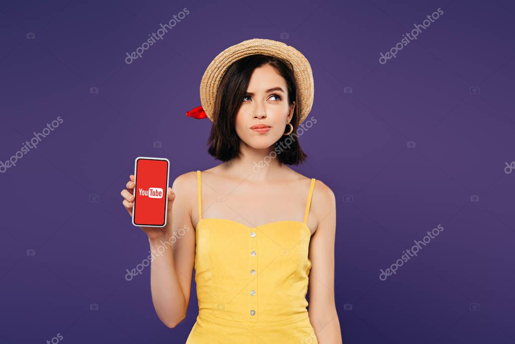 KYIV, UKRAINE - JULY 3, 2019: dreamy pretty girl in straw hat holding smartphone with youtube app isolated on purple