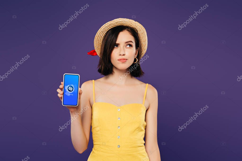 KYIV, UKRAINE - JULY 3, 2019: dreamy pretty girl in straw hat holding smartphone with shazam app isolated on purple