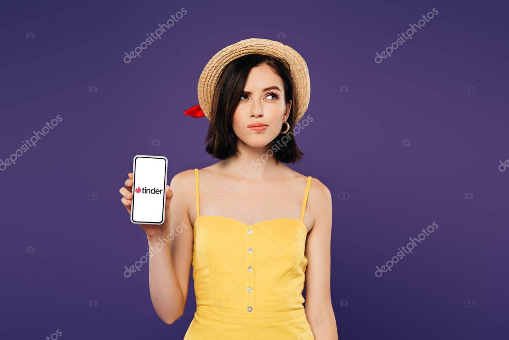 KYIV, UKRAINE - JULY 3, 2019: dreamy pretty girl in straw hat holding smartphone with tinder app isolated on purple