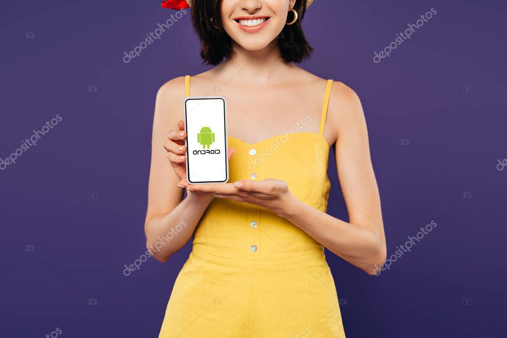 KYIV, UKRAINE - JULY 3, 2019: cropped view of smiling girl holding smartphone with android logo isolated on purple