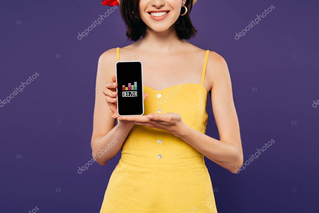 KYIV, UKRAINE - JULY 3, 2019: cropped view of smiling girl holding smartphone with deezer app isolated on purple