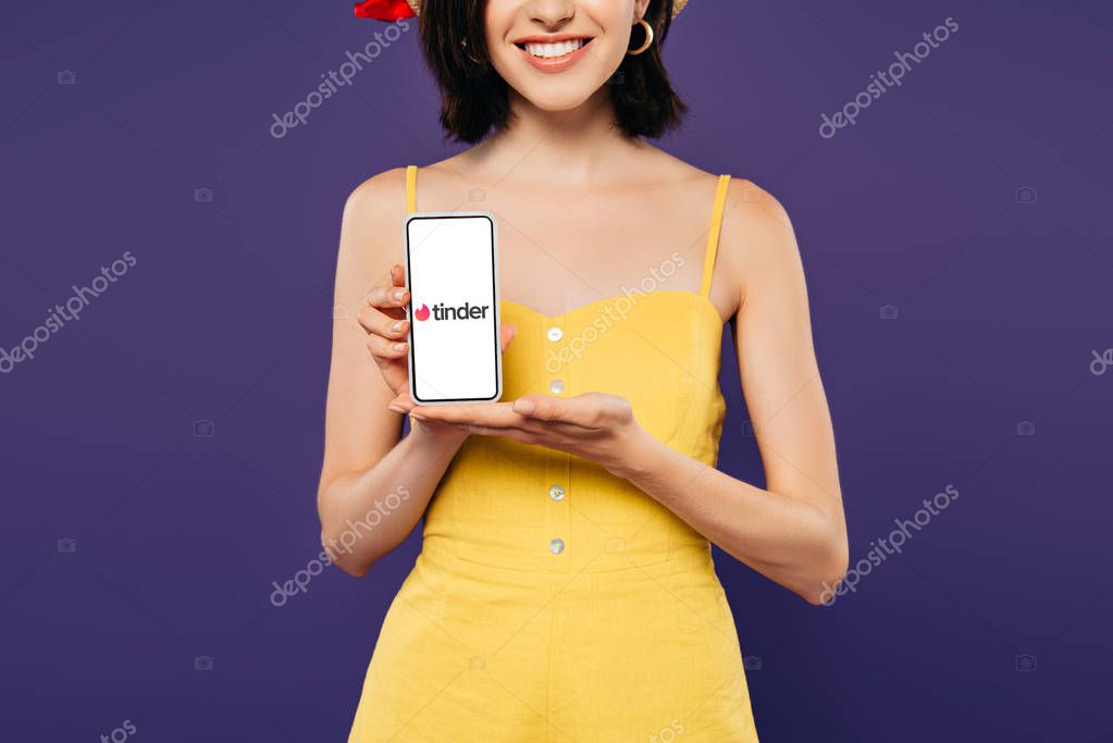 KYIV, UKRAINE - JULY 3, 2019: cropped view of smiling girl holding smartphone with tinder app isolated on purple