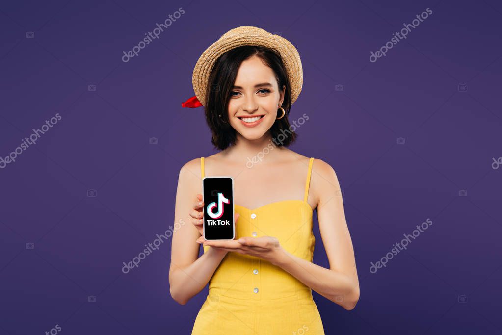 KYIV, UKRAINE - JULY 3, 2019: smiling pretty girl in straw hat holding smartphone with tiktok app isolated on purple