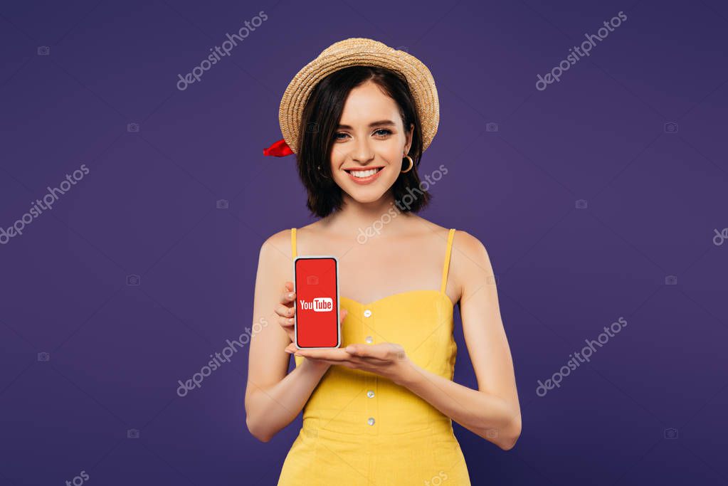 KYIV, UKRAINE - JULY 3, 2019: smiling pretty girl in straw hat holding smartphone with youtube app isolated on purple