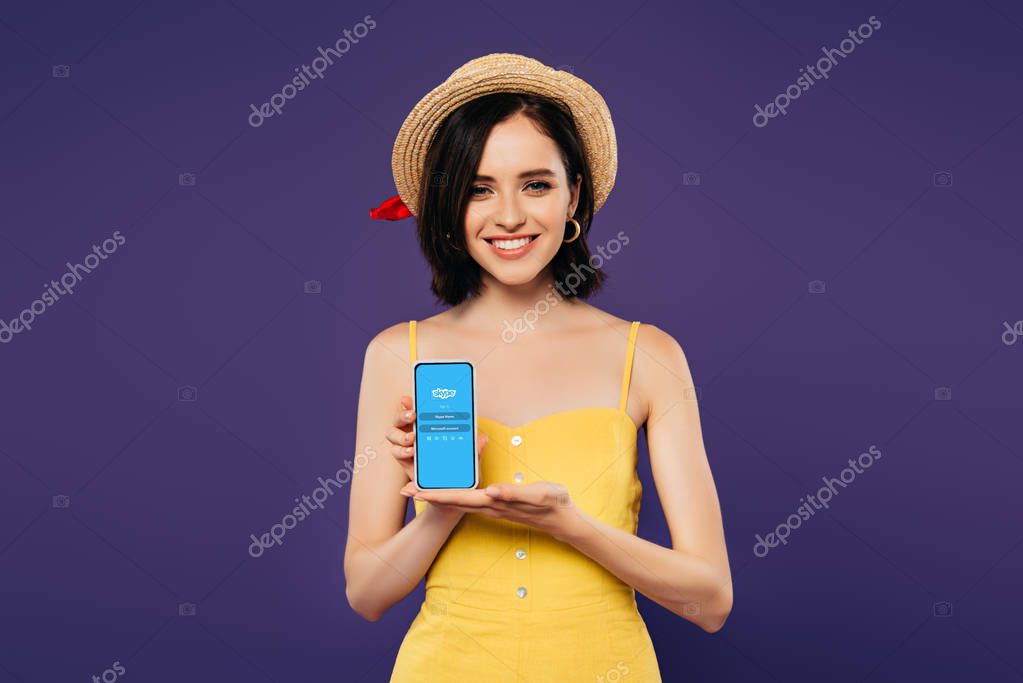 KYIV, UKRAINE - JULY 3, 2019: smiling pretty girl in straw hat holding smartphone with skype app isolated on purple