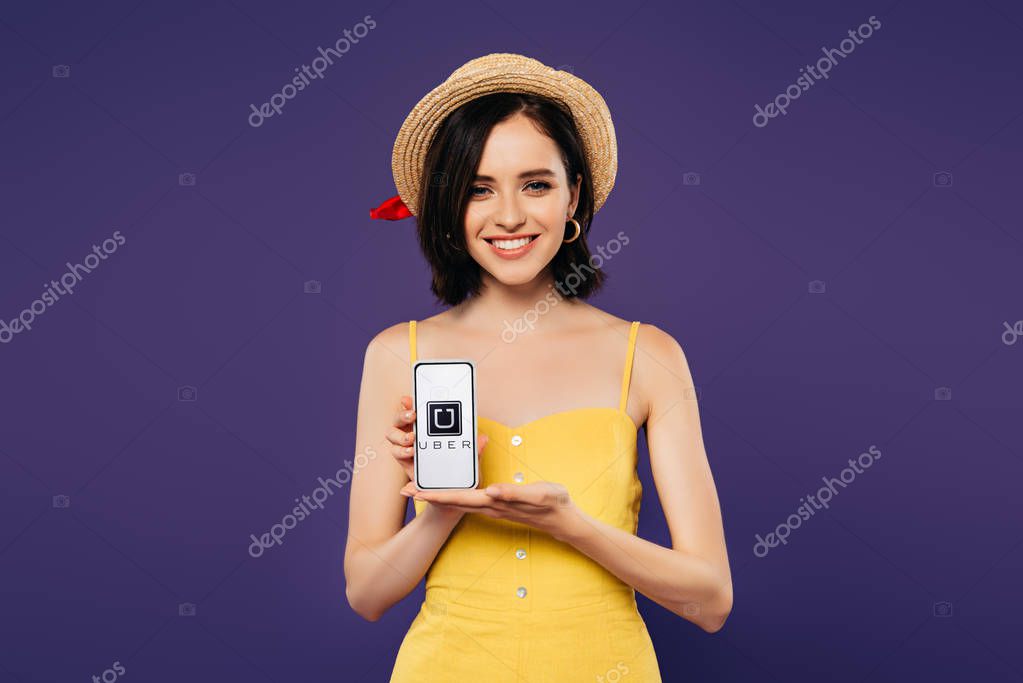 KYIV, UKRAINE - JULY 3, 2019: smiling pretty girl in straw hat holding smartphone with uber app isolated on purple