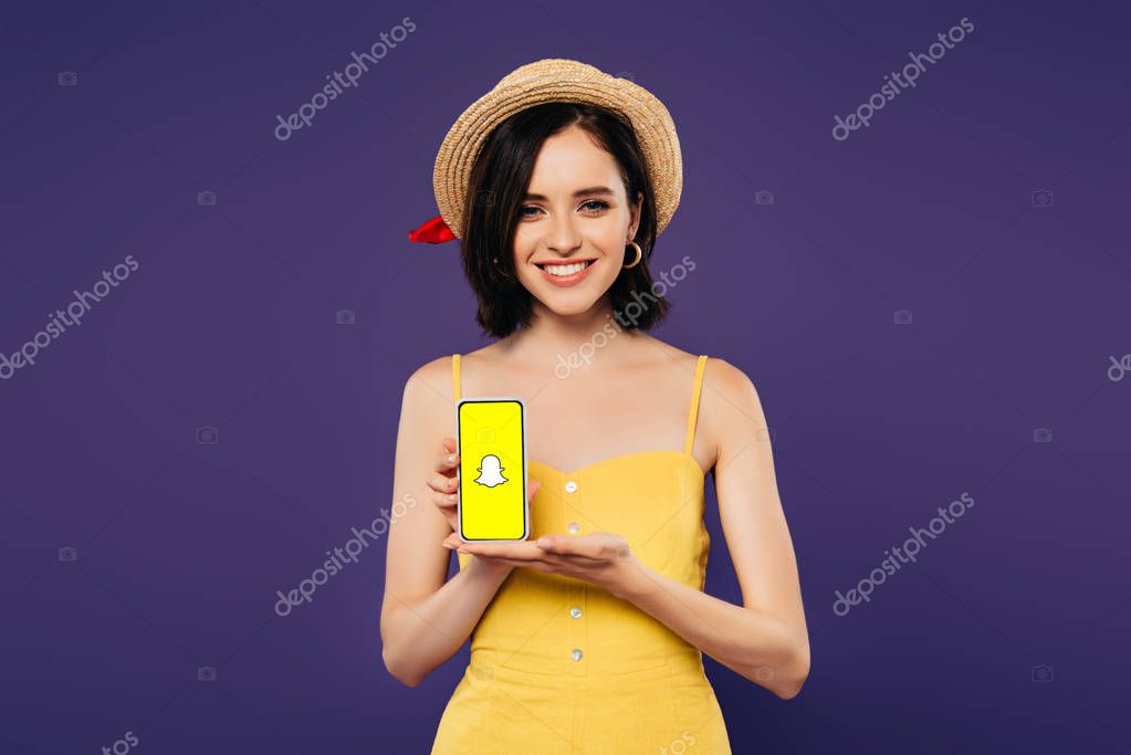 KYIV, UKRAINE - JULY 3, 2019: smiling pretty girl in straw hat holding smartphone with snapchat app isolated on purple