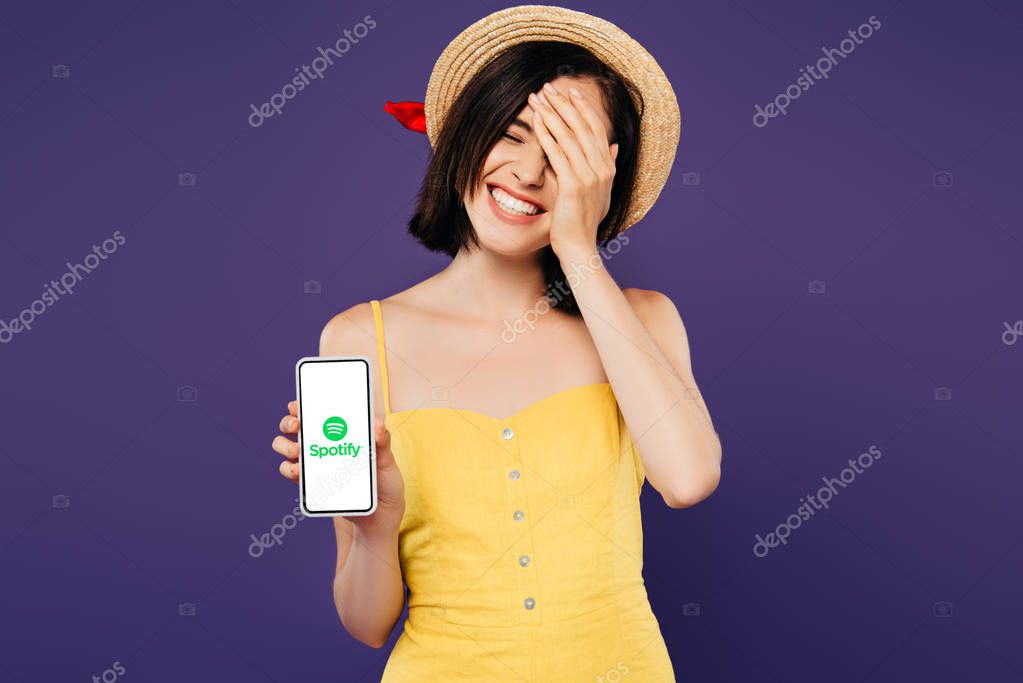 KYIV, UKRAINE - JULY 3, 2019: smiling pretty girl in straw hat with hand on face showing smartphone with spotify app isolated on purple