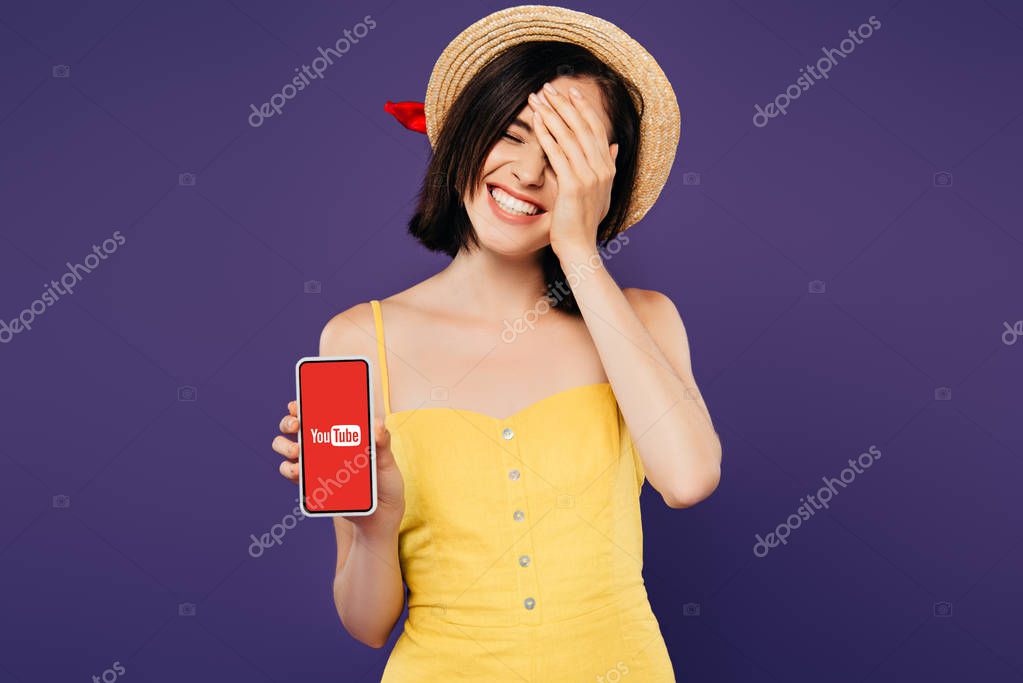KYIV, UKRAINE - JULY 3, 2019: smiling pretty girl in straw hat with hand on face showing smartphone with youtube app isolated on purple