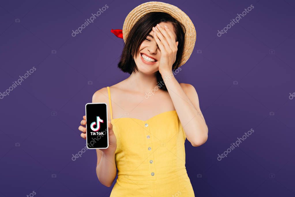 KYIV, UKRAINE - JULY 3, 2019: smiling pretty girl in straw hat with hand on face showing smartphone with tiktok app isolated on purple