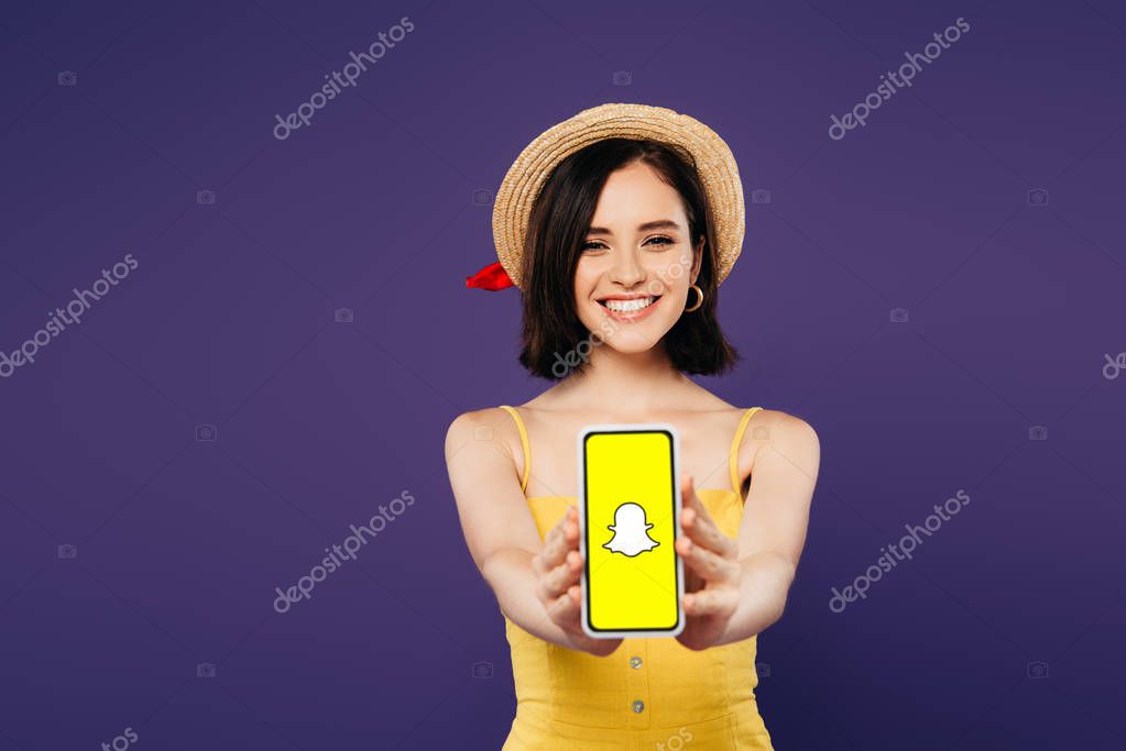 KYIV, UKRAINE - JULY 3, 2019: selective focus of smiling pretty girl in straw hat presenting smartphone with snapchat app isolated on purple