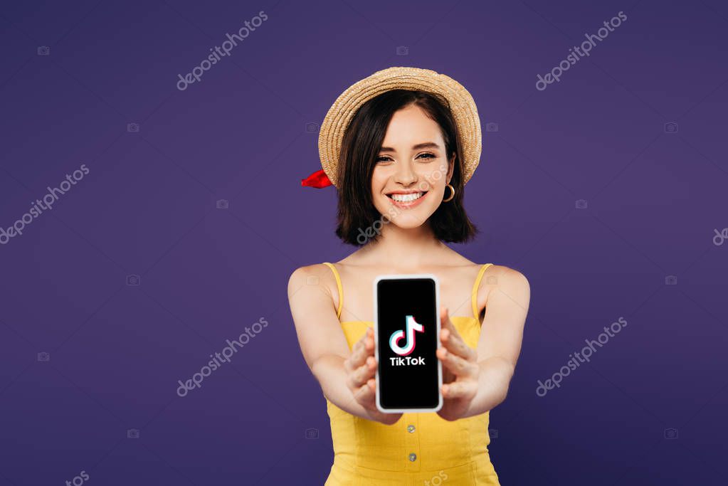 KYIV, UKRAINE - JULY 3, 2019: selective focus of smiling pretty girl in straw hat presenting smartphone with tiktok app isolated on purple
