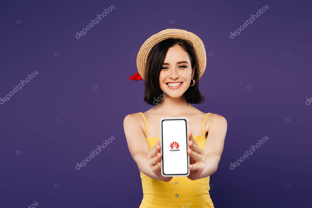 KYIV, UKRAINE - JULY 3, 2019: selective focus of smiling pretty girl in straw hat presenting smartphone with huawei logo isolated on purple