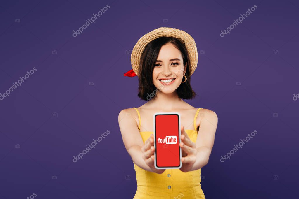 KYIV, UKRAINE - JULY 3, 2019: selective focus of smiling pretty girl in straw hat presenting smartphone with youtube app isolated on purple