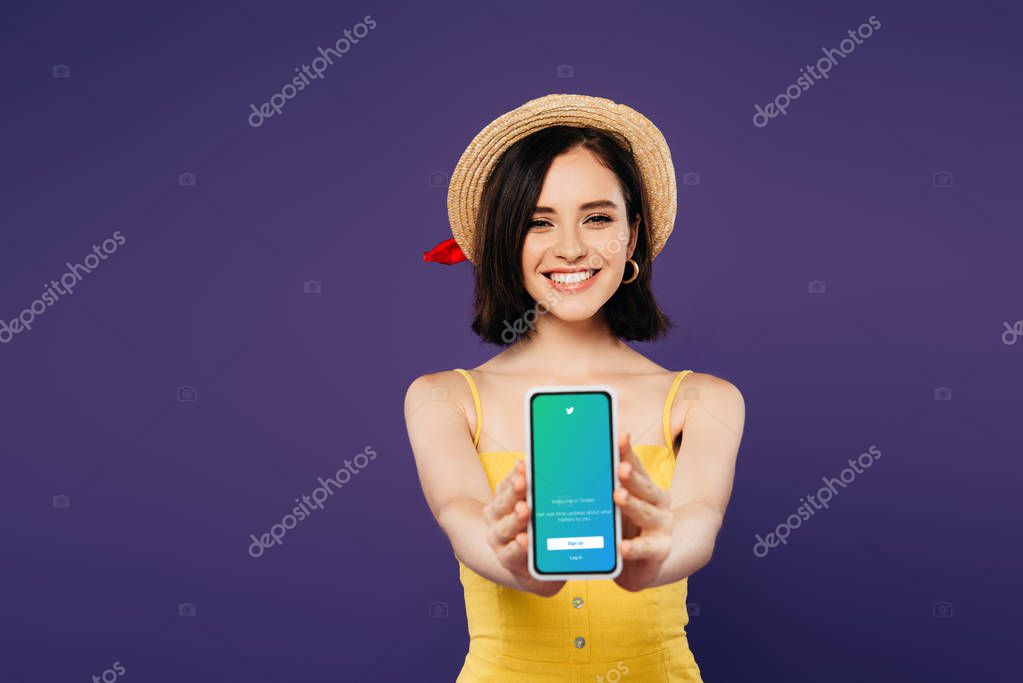 KYIV, UKRAINE - JULY 3, 2019: selective focus of smiling pretty girl in straw hat presenting smartphone with twitter app isolated on purple
