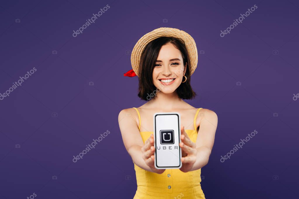 KYIV, UKRAINE - JULY 3, 2019: selective focus of smiling pretty girl in straw hat presenting smartphone with uber app isolated on purple