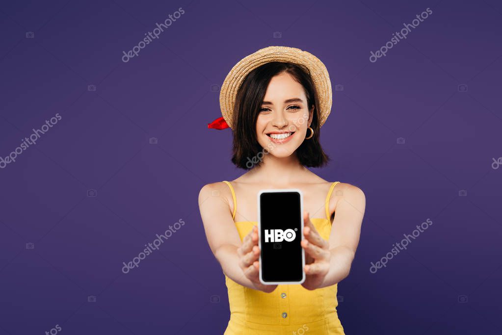 KYIV, UKRAINE - JULY 3, 2019: selective focus of smiling pretty girl in straw hat presenting smartphone with hbo app isolated on purple