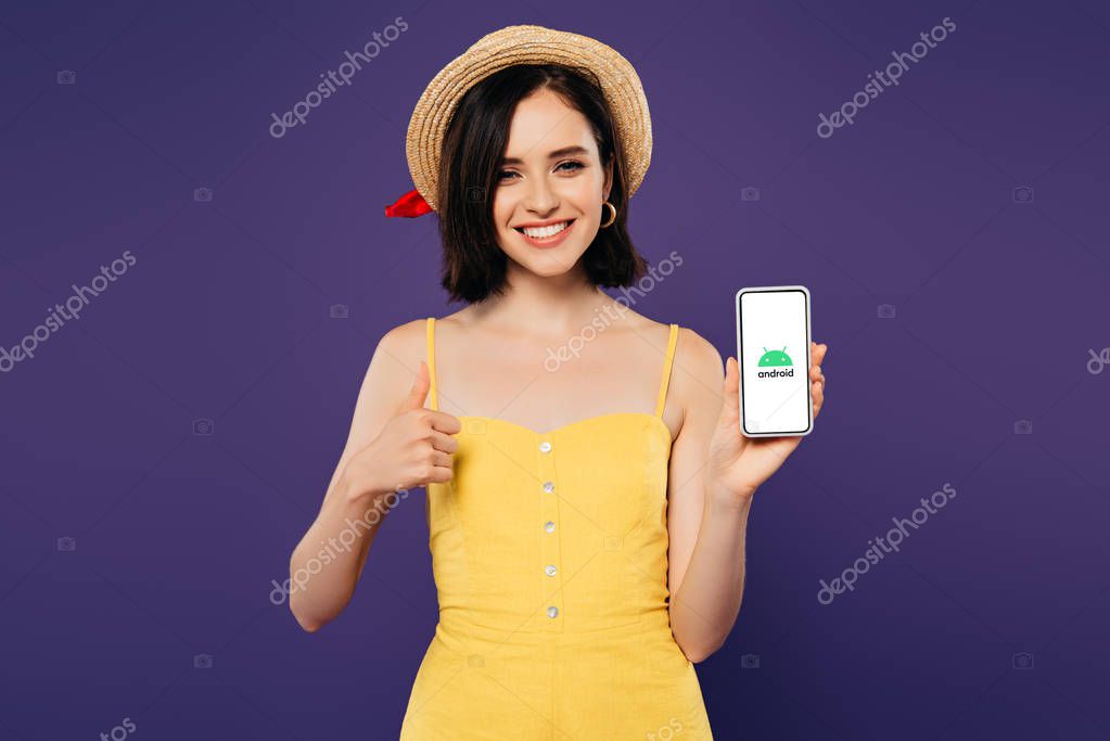 KYIV, UKRAINE - JULY 3, 2019: smiling pretty girl in straw hat showing thumb up while holding smartphone with android logo isolated on purple
