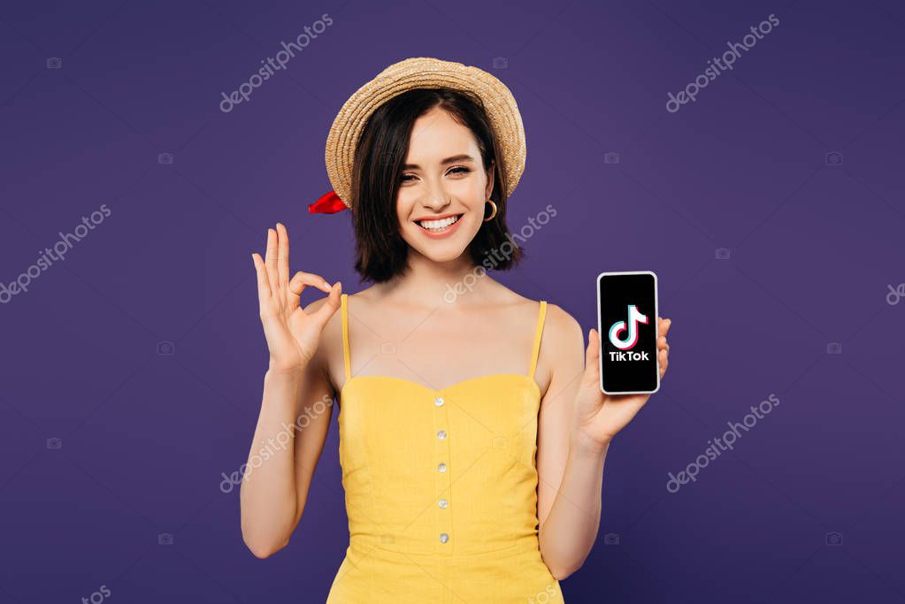 KYIV, UKRAINE - JULY 3, 2019: smiling pretty girl in straw hat holding smartphone with tiktok app and showing ok sign isolated on purple