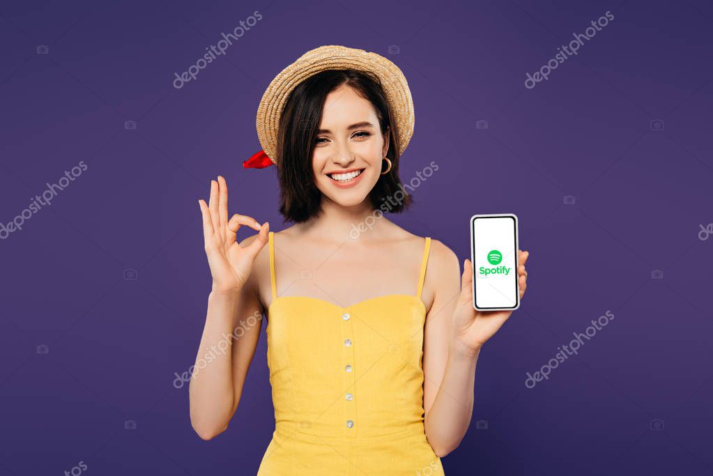 KYIV, UKRAINE - JULY 3, 2019: smiling pretty girl in straw hat holding smartphone with spotify app and showing ok sign isolated on purple