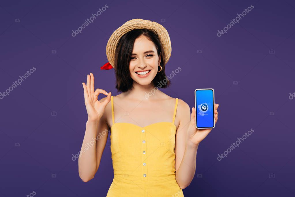 KYIV, UKRAINE - JULY 3, 2019: smiling pretty girl in straw hat holding smartphone with Shazam app and showing ok sign isolated on purple