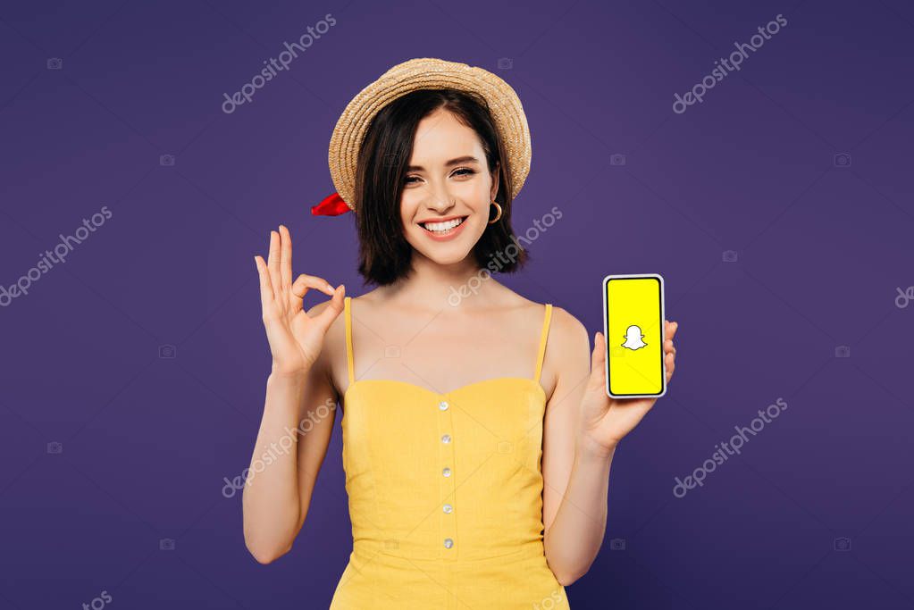 KYIV, UKRAINE - JULY 3, 2019: smiling pretty girl in straw hat holding smartphone with Snapchat app and showing ok sign isolated on purple