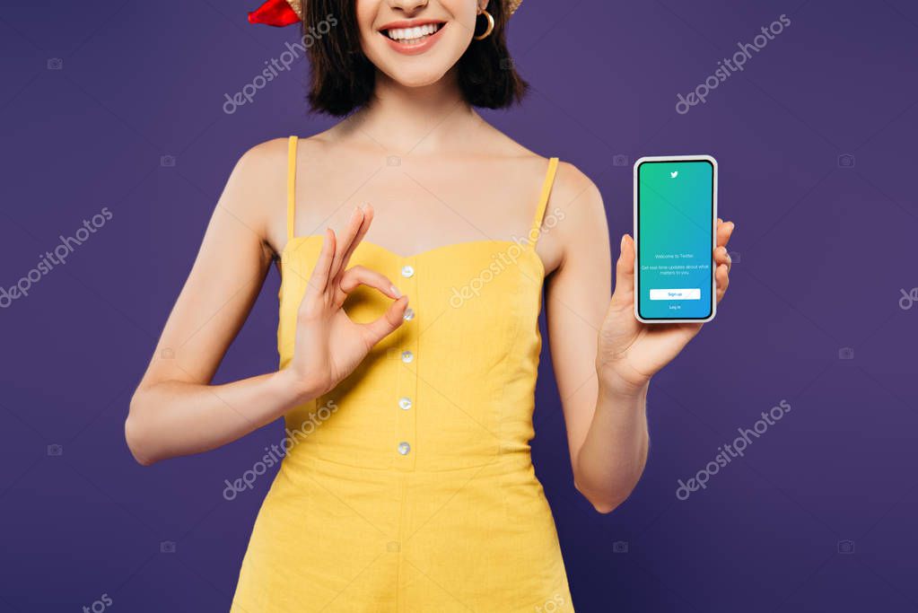 KYIV, UKRAINE - JULY 3, 2019: cropped view of smiling girl in holding smartphone with twitter logo and showing ok sign isolated on purple