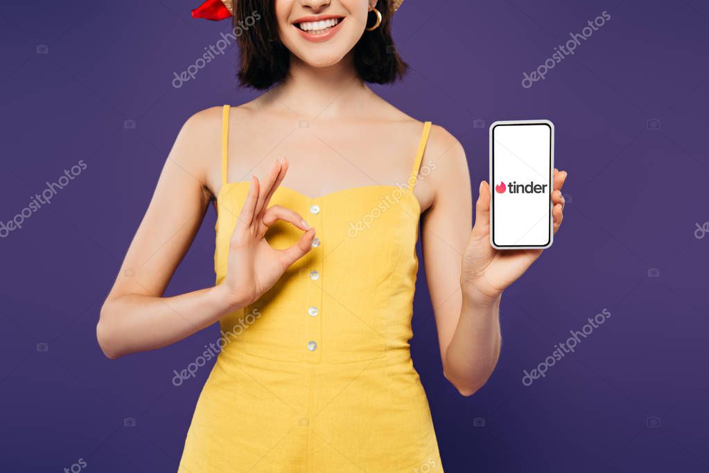 KYIV, UKRAINE - JULY 3, 2019: cropped view of smiling girl in holding smartphone with tinder logo and showing ok sign isolated on purple