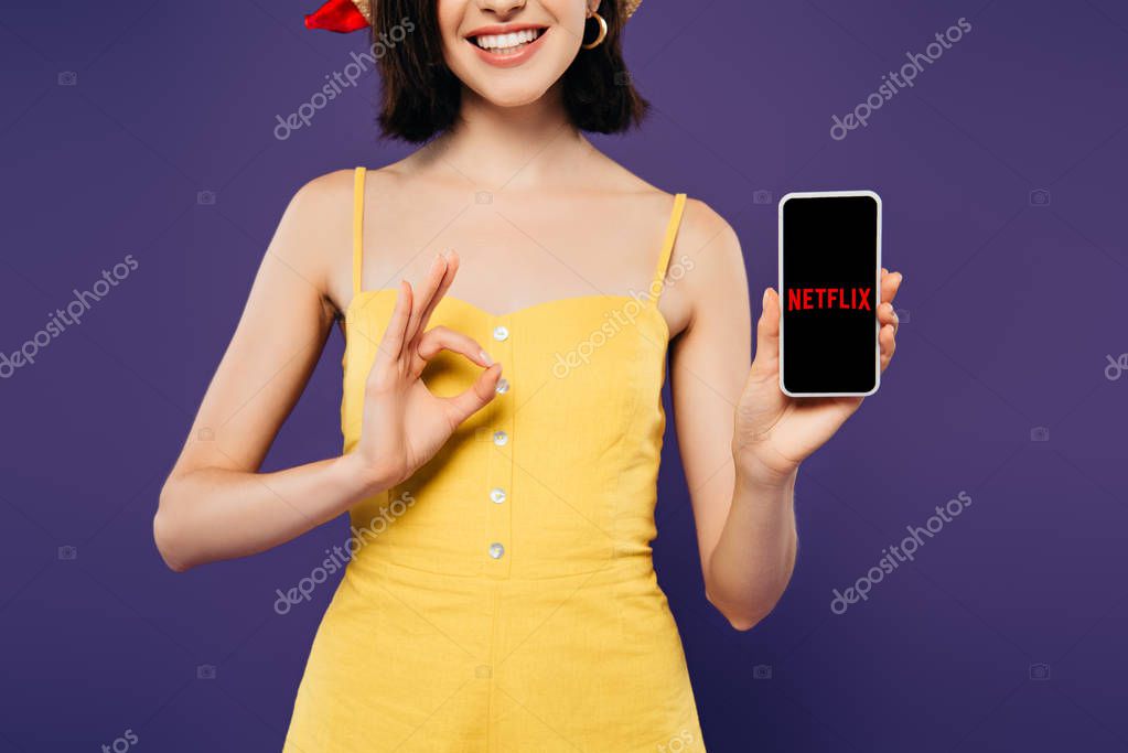 KYIV, UKRAINE - JULY 3, 2019: cropped view of smiling girl in holding smartphone with netflix logo and showing ok sign isolated on purple