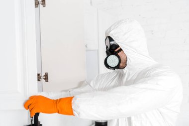 exterminator in protective mask and uniform standing near kitchen cabinet  clipart