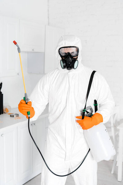 exterminator in protective mask and uniform standing with toxic spray in kitchen 